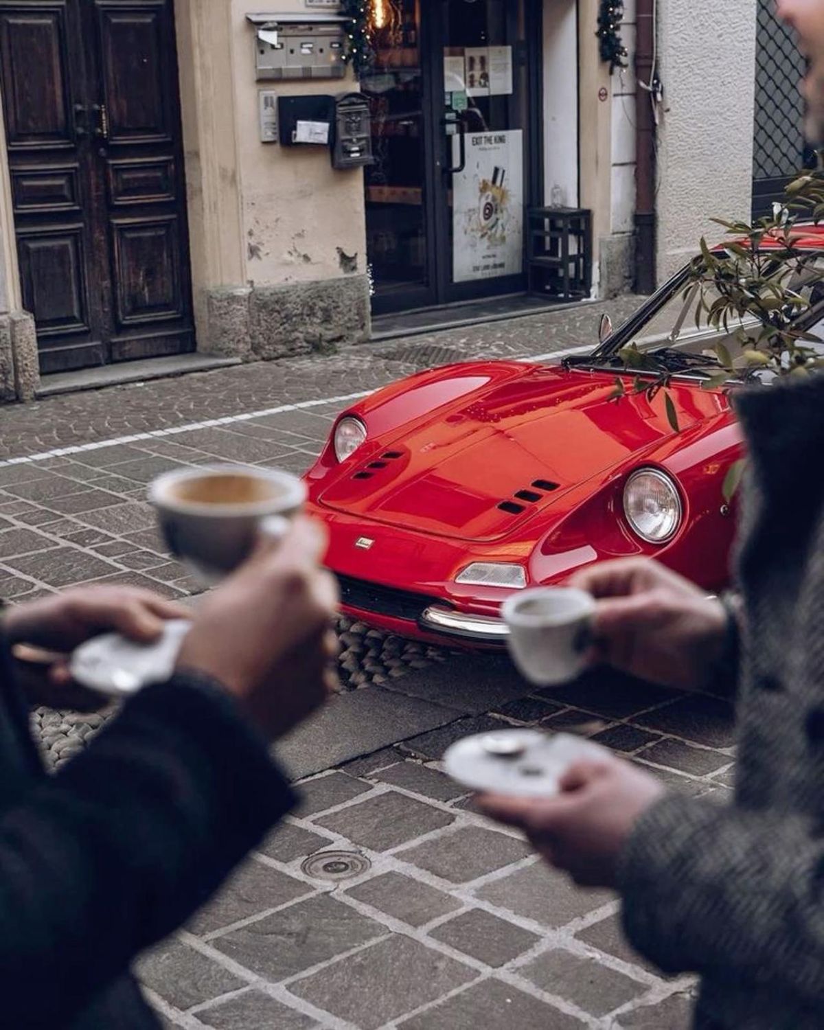 Two people holding coffee cups in front of a red sports car on a street.