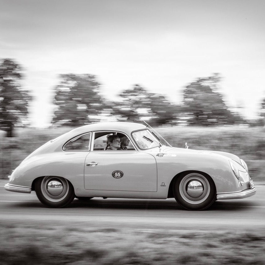 A vintage sports car photographed in side profile on a road with blurred trees in the background, rendered in black and white.