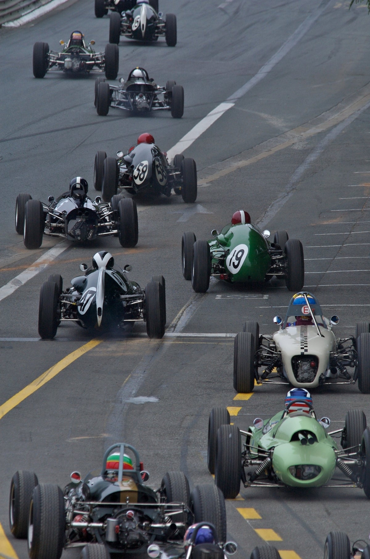 A high-angle action shot of multiple vintage open-wheel race cars racing on a paved track, with drivers wearing helmets and goggles.